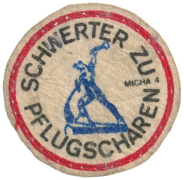 Das Bild zeigt einen Aufnäher mit der schwarzen kreisförmigen Aufschrift "Schwerter zu Pflugscharen" umrahmt mit einem roten Kreis, darin in blau ein Abbild der gleichnamigen Skulptur