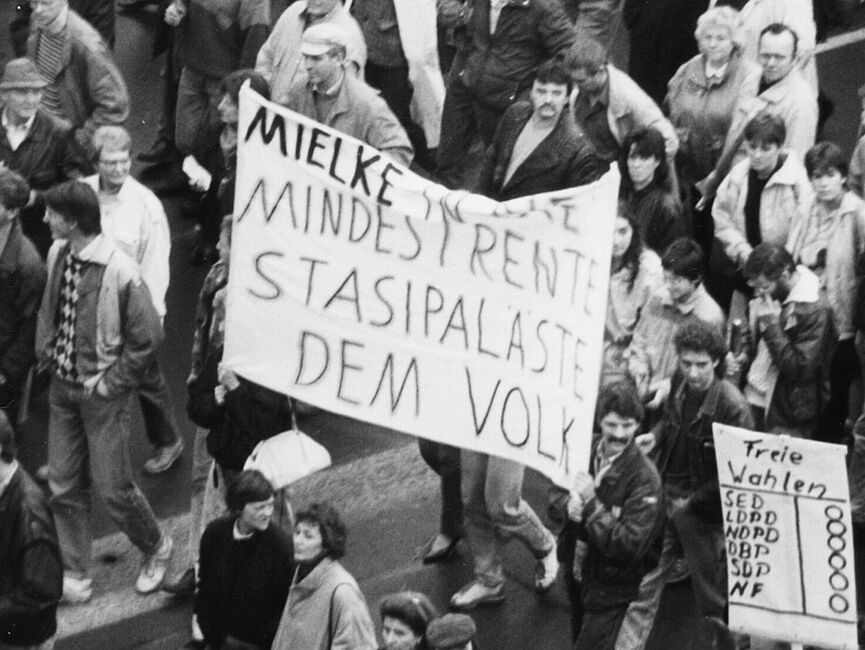 Foto einer Demonstration. In der Bildmitte steht auf einem Transparent "Mielke in die Mindestrente Stasipaläste dem Volk", auf einem rechts daneben ist ein Wahlzettel gemalt, darüber steht "Freie Wahlen".
