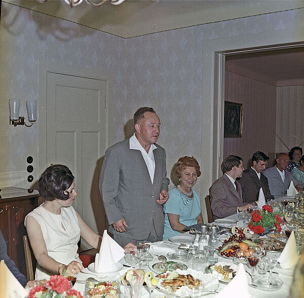 Eine festlich gedeckte Tafel in einem kleinen Wohnzimmer. Am Tsich sitzen Gäste, Erich Mielke im Freizeitanzug steht und spricht.