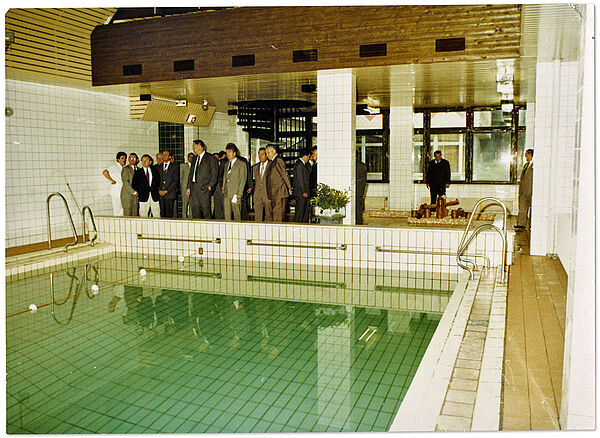 Blick in eine kleine Schwimmhalle in einem neuen Stasi-Hotel über den Pool hinweg auf eine Gruppe von Herren, die sich die Anlage anlässlich der Einweihung des Hotelgebäudes ansehen.