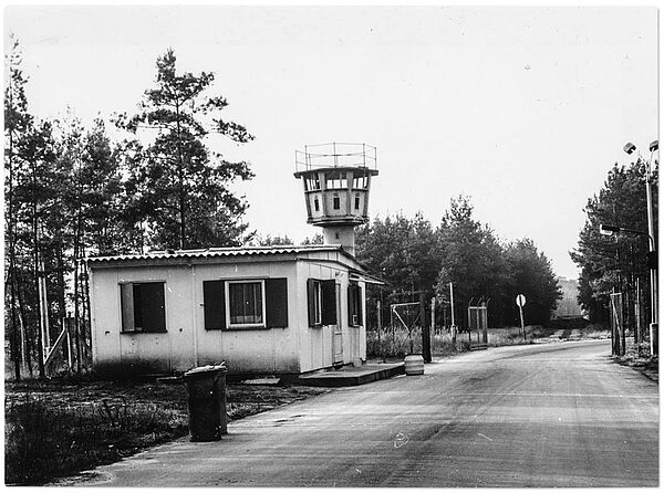 Blick die Zufahrtsstraße zum Stasi-Gelände in Freienbrink hinunter: links der Straße ein hüttenartiges Wärterhäuschen, dahinter ein Wachturm.