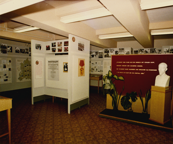 Ein Ausstellungsraum: An den Wänden und einer Stellwand im Raum sind Bilder und Texte angebracht. Rechts im Mittelgrund eine halbhohe rote Wand mit der Aufschrift: "Tschekist sein kann nur ein Mensch mit kühlem Kopf, heißem Herzen und sauberen Händen. Ein Tschekist muss sauberer und ehrlicher als irgendwer – er muss so klar wie ein Kristall sein". Rechts daneben steht eine Büste von Feliks Dzierżyński. 