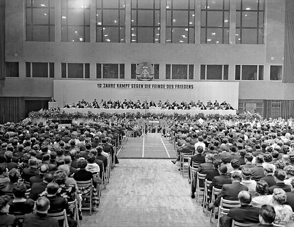 Blick durch eine schlichte, große Halle mit zahlreichen auf Stühlen sitzenden Zuschauern. Ihre Stühle sind auf ein großes, mit zahlreichen Personen besetztes Podium gerichtet. Hinter dem Podium die Losung: „10 Jahre Kampf gegen die Feinde des Friedens“. Darüber das Wappen der DDR.