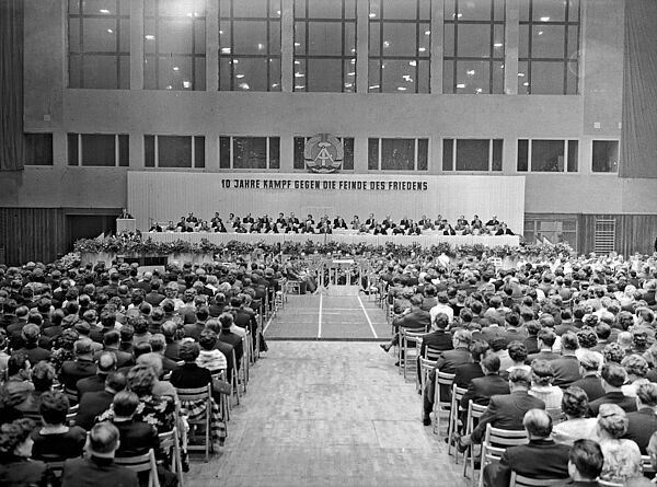 Blick durch eine schlichte, große Halle mit zahlreichen auf Stühlen sitzenden Zuschauern. Ihre Stühle sind auf ein großes, mit zahlreichen Personen besetztes Podium gerichtet. Hinter dem Podium die Losung: „10 Jahre Kampf gegen die Feinde des Friedens“. Darüber das Wappen der DDR.