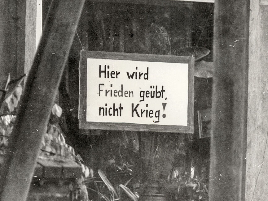 Schild am Wohnzimmerfenster der Pfarrei mit dem Text:"Hier wird Frieden geübt, nicht Krieg!" Schwarz-weiß Foto aus der Bilddokumentation der Stasi: handgemaltes Schild hängt an Schnüren im Wohnzimmerfenster der Pfarrei mit dem Text: "Hier wird Frieden geübt, nicht Krieg!"