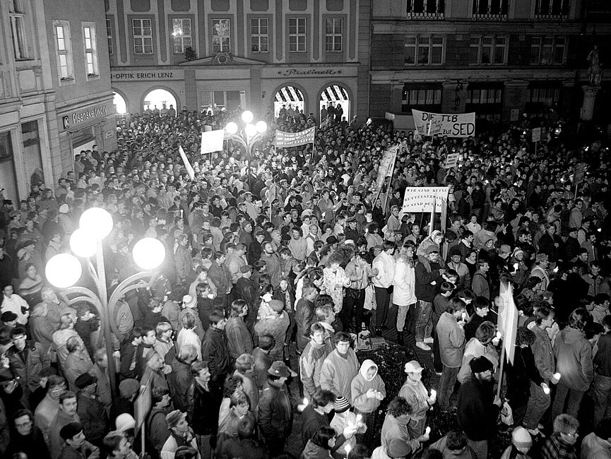 Foto einer Menschenmenge, die sich Abends mit Transparenten auf einem Platz versammelt.