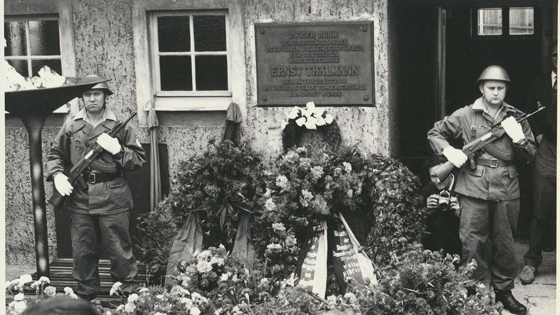 Schwarz-weiß Foto: Zwei uniformierte und bewaffnete Soldaten stehen in einem Hof, zwischen ihnen eine Plakette, die an die dort stattgefundene Ermordung Ernst Thälmanns erinnert, vor und neben ihnen eine Vielzahl von Blumen