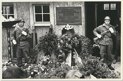 Zwei uniformierte und bewaffnete Soldaten stehen in einem Hof neben einer Plakette, die an die dort stattgefundene Ermordung Ernst Thälmanns erinnert. Vor und neben ihnen ist eine Vielzahl von Blumengestecken und Blumenkränzen.