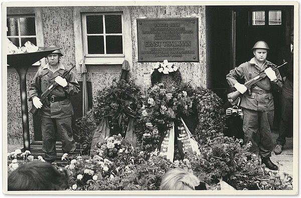 Schwarz-weiß Foto: Zwei uniformierte und bewaffnete Soldaten stehen in einem Hof, zwischen ihnen eine Plakette, die an die dort stattgefundene Ermordung Ernst Thälmanns erinnert, vor und neben ihnen eine Vielzahl von Blumen