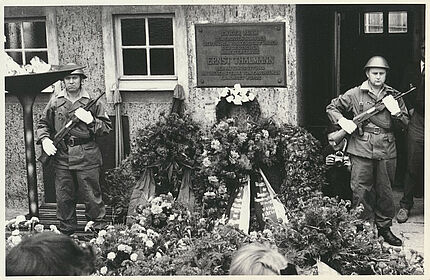 Schwarz-weiß Foto: Zwei uniformierte und bewaffnete Soldaten stehen in einem Hof, zwischen ihnen eine Plakette, die an die dort stattgefundene Ermordung Ernst Thälmanns erinnert, vor und neben ihnen eine Vielzahl von Blumen