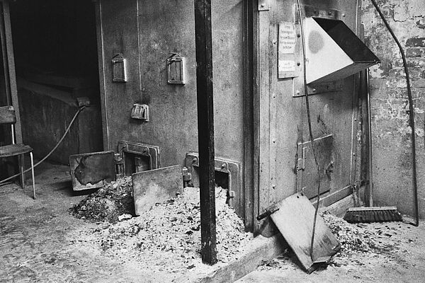 Raum, in dem sich zwei Öfen befinden, vor denen eine große Menge verbranntes Papier liegt.