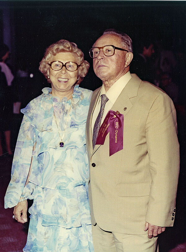 Ein älteres Ehepaar in festlicher Kleidung steht eng beieinander, sie lächelt strahlend, er blickt streng.