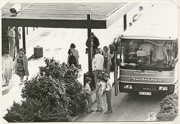 Die Stasi beobachtete die Reisegruppe der Landeszentrale für politische Bildung Schleswig-Holstein vor dem Rostocker Interhotel "Warnow", 15. August 1988; Quelle: BArch, MfS, BV Rostock, Abt. VI, Nr. 11, Teil 2, Bl. 208, Bild 2 Schwarz-Weiß-Foto, das einen überdachten Eingangsbereich zu einem Gebäude zeigt. Davor befinden sich diverse Personengruppen, im vorderen Bildteil stehen drei Kinder neben einem Reisebus. Daneben stehen Menschen mit Koffern.