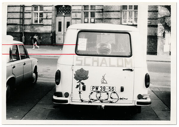 Foto eines weißen Kleinbusses auf dem "Schalom" steht, der Aufkleber einer Rose, deren Stiel aus Stacheldraht ist, sowie das "Schwerter zu Pflugscharen"-Symbol klebt. Auf dem Schwazweissfoto sind zwei rote waagerechte Striche an Aufklebern des Autos von der Stasi eingezeichnet.