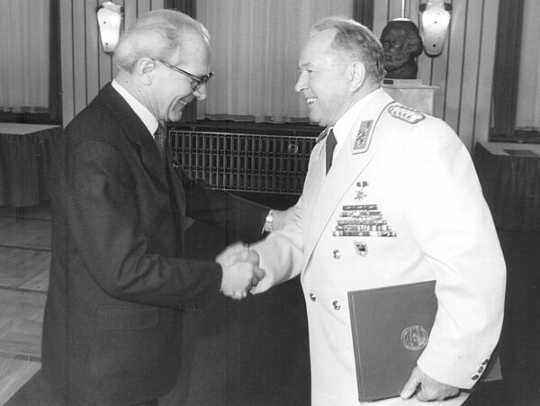 Honecker und Mielke schüttel sich freudig lächelnd die Hände. Honecker trägt einen schwarzen Anzug, Mielke eine weiße Galauniform.