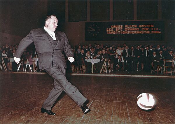 Im Innern einer Sporthalle. Im Hintergund festlich gedeckte Tafeln, daran sitzen und stehen festlich gekleidete Menschen. rechts über dieser Szene eine Anzeigetafel: „Gruß allen Gästen des BFC Dynamo zur III. Traditionsveranstaltung“. Im Vordergrund Erich Mielke, der gerade mit angespannten Gesichtszügen einen Fußball geschossen hat.