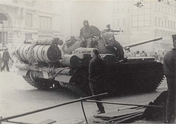 Soldaten sitzen auf einem Panzer, der auf einer Straße steht. Im Hintergrund ist eine Menschenmenge zu sehen.