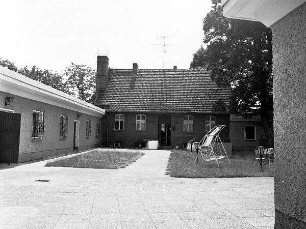 Blick in einen Hof. Im Hintergrund ein aus roten Ziegeln errichtetes Wohnhaus. Im Vordergrund eine mit Platten gepflasterte Fläche. Dazwischen eine Rasenfläche auf der eine Hollywood-Schaukel steht. links davon ein eingeschossiger Neubau-Gebäudetrakt. Rechts im Vordergrund ist ein Teil der Außenwand eines weiteren Gebäudes zu sehen.