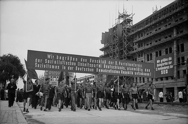 Schwarz-Weiß-Foto einer in FDJ-Uniformen gekleideten Menschenmenge mit Fahnen und einem Hochtransparent. Auf diesem steht "Wir begrüßen den Beschluß der II. Parteikonferenz der Sozialistischen Einheitspartei Deutschlands, planmäßig den Sozialismus in der Deutschen Demokratischen Republik aufzubauen!". Rechts am Straßenrand ist ein im Bau befindliches Gebäude mit einem Transparent mit der Aufschrift "Wir grüßen die Delegierten der II. Parteikonferenz der SED"