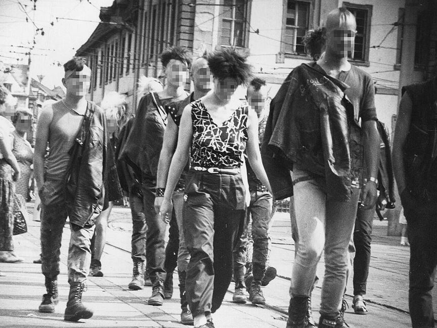 Schwarz-Weiß-Foto einer Gruppe Jugendlicher im Punk-Look auf einem Platz mit Straßenbahnschienen. Ältere Damen drehen sich nach ihnen um. Oben ein Netz aus Oberleitungen, im Hintergrund Häuser.