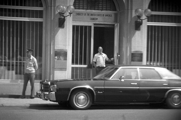 Die Stasi überwachte den Eingang der US-Botschaft an der Neustädtischen Kirchstraße in Ost-Berlin, Ende der 80er Jahre; Quelle: BArch, MfS, HA II, Fo, Nr. 1009, Bild 25 Schwarzweißfoto, zu sehen ist eine Person die aus einem Gebäude auf die Straße tritt. Vor ihr steht ein dunkles Auto. Rechts im Bild, an der Fassade des Gebäudes, steht eine Person die Uniform trägt.