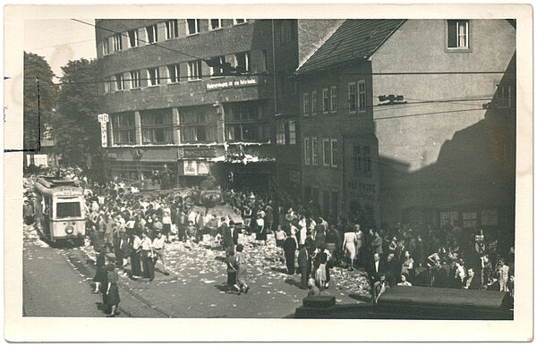 Schwarz-Weiß-Foto eines Platzes. Zu sehen ist eine kleinere Menschenmenge, die einem Panzer umringt. Am rechten Rand ist eine Straßenbahn zu sehen.