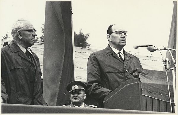 Blick von auf ein Podium. Ein Redner am Pult, hinter ihm ist der Kopf von Martin Weikert zu erkennen.