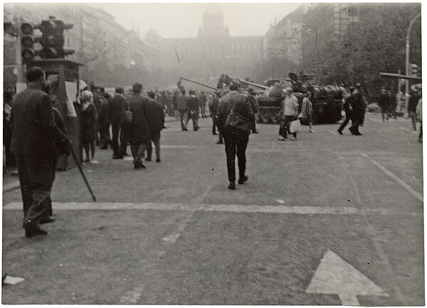 Schwarzweißbild Prag 1968: auf einem Platz inmittewn von kreuz und quer gehenden Menschen steht ein sowjetischer Panzer. Die Geschützrohre weiterer Panzer ragen von rechts in das Bild und aus der Menschenmenger heraus.