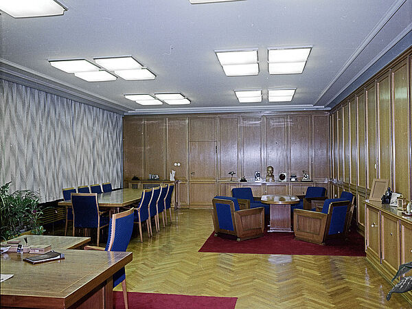 Blick in einen großen Büroraum: vorne links im Vordergrund die rechte Ecke eines Schreibtischs, hinten rechts eine Sitzgruppe mit Tisch und Sesseln, links ein Konferenztisch mit Stühlen. Alle Sitzmöbel haben blaue Bezüge.