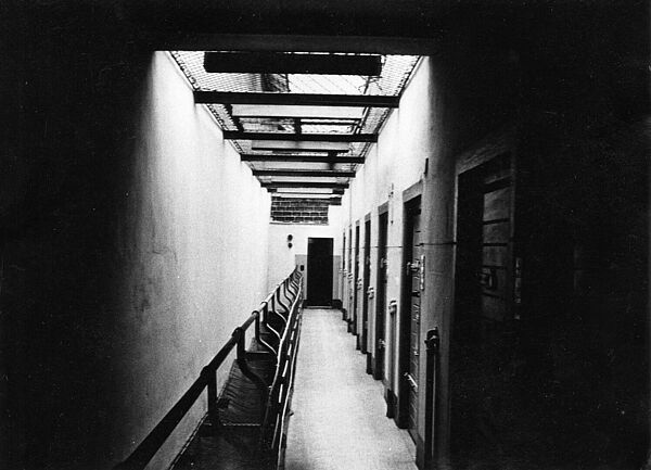 Gefängnistrakt in der Zentralen Untersuchungshaftanstalt der Stasi in Berlin-Lichtenberg, Magdalenenstraße, um 1990; Quelle: Stefan Eikermann Schwarz-Weiß-Foto eines Gefängnistrakts. AUf der rechten Seite befinden sich Zellentüren, die offene Decke des Ganges ist mit einem Netz verhangen.
