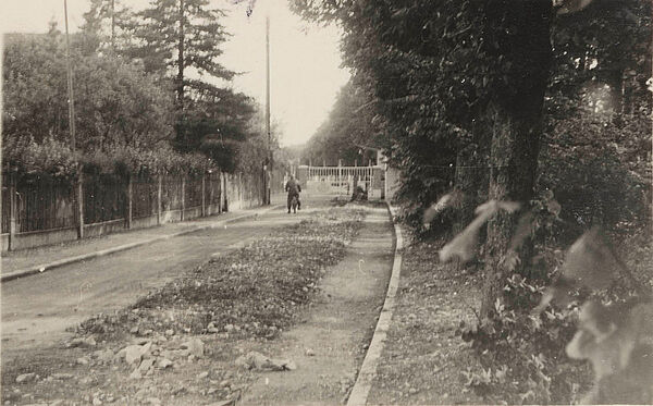 Einfahrt zum Sitz der Organisation Gehlen in Pullach, Juli 1953 Blick eine Schotter-Straße in schlechtem Zustand hinunter. Sie ist von beiden Seiten von Zäunen umgeben. Im Hintergrund ist ein geschlossenes Einfahrtstor zu sehen.