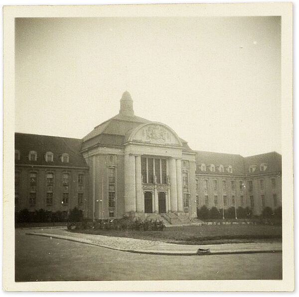 Bezirksverwaltung Schwerin des Ministeriums für Staatssicherheit, Demmlerplatz 1-2; Quelle: BArch, MfS, BV Schwerin, AU, Nr. 42/55, Bd. 1, Bl. 122 Blick über den Demmlerplatz aufSchwarz-weiß Foto: Blick über einen Platz mit Rasenfläche in der Mitte, dahinter ein breites 2-stöckiges Gebäude mit Dachgeschoss, in der Mitte des Gebäudes ein großer Vorbau mit Säulen und Türmchen auf dem Dach – hier befindet sich der Eingang.
