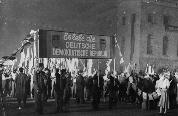 Das Foto zeigt eine Demonstration, in der Bildmitte sind mehrere Männer zu sehen, die ein Transparent an Stöcken hochhalten, auf dem das Logo der "FDJ" zwei Mal abgebildet ist und der Text steht: "Es lebe die Deutsche Demokratische Republik". Dahinter stehen zahlreiche Menschen mit Fahnen.