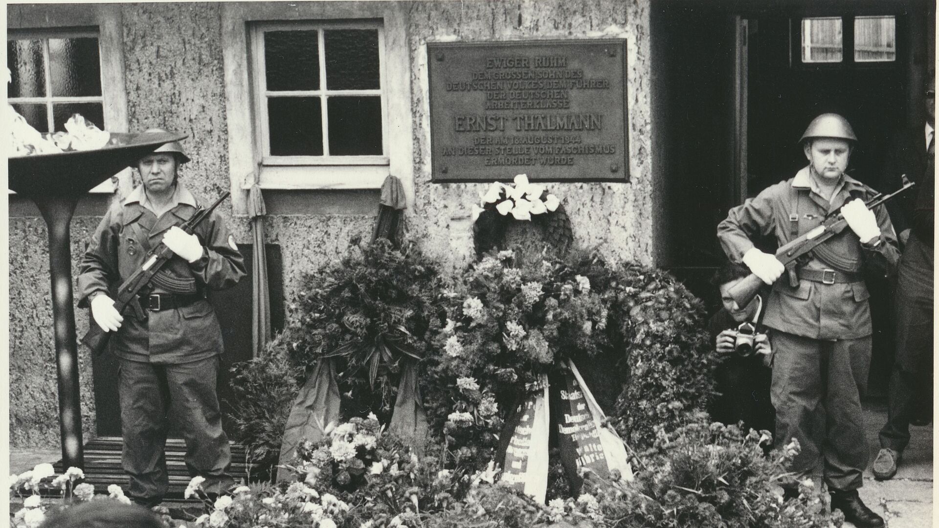 Zwei uniformierte und bewaffnete Soldaten stehen in einem Hof neben einer Plakette, die an die dort stattgefundene Ermordung Ernst Thälmanns erinnert. Vor und neben ihnen ist eine Vielzahl von Blumengestecken und Blumenkränzen.