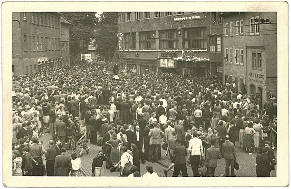 Schwarz-Weiß-Foto einer großen Menschenmenge auf einem Platz, der von Gebäuden umgeben ist.