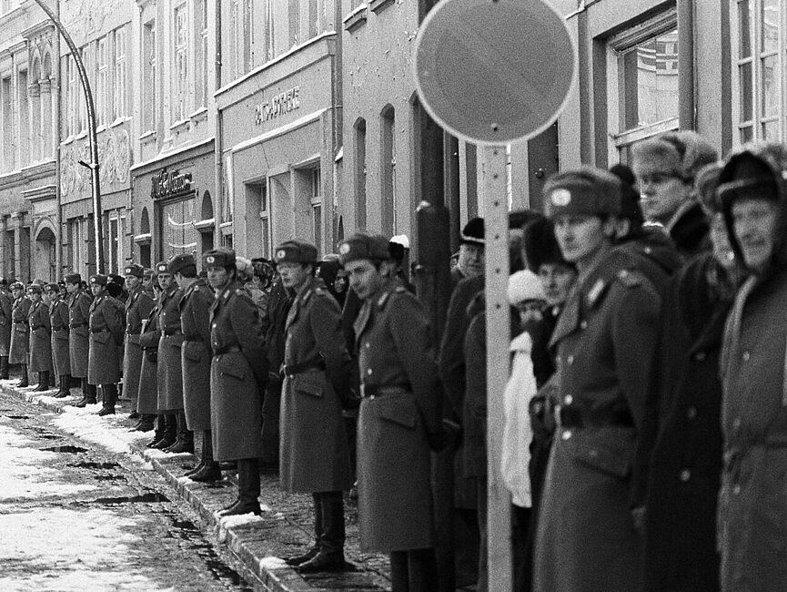 Blick eine nach links gekurvte Straße hinunter. Nur der rechte Straßenrand ist zu sehen, dahinter Gebäude. Am Straßenrand stehen Schulter an Schulter uniformierte Volkspolizisten Spalier, dahinter einige Schaulustige.