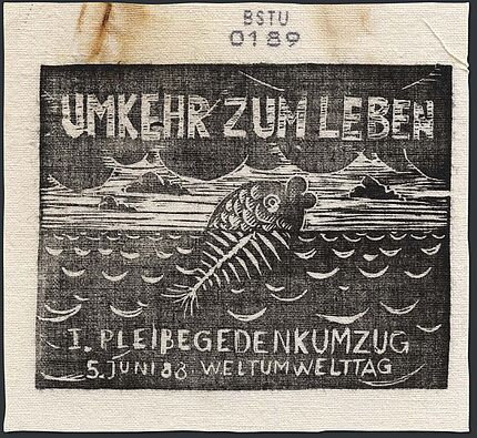 Bedrucktes Stoffstück, auf dem ein Fisch abgebildet ist, der aus dem Wasser herausschaut. Der untere, sich im Wasser befindliche Teil des Fisches besthet nur aus Gräten. Darüber steht der Text "Umkehr zum Leben", darunter "1. Pleißegedenkumzug, 5. Juni '88, Weltumwelttag