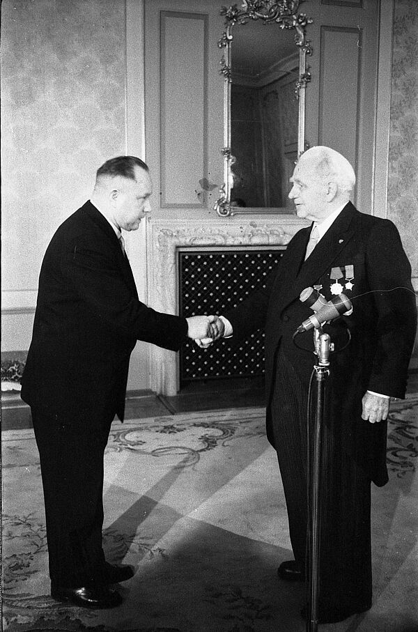 Im Saal eines Schlosses schütteln sich zwei Männer in festlicher Kleidung die Hand.