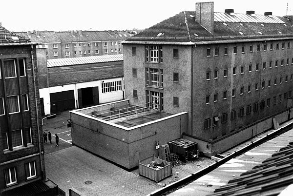Gebäude der ehemaligen Untersuchungshaftanstalt der Stasi in Rostock, 1990 Schwarz-Weiß-Foto. Das Bild zeigt den Blick in den Innenhof eines Gebäudekomplexes. In der Mitte ist ein Vorbei der zu einem vierstöckigen Gebäude gehört, dessen Fenster zum Teil vergittert sind. Links neben dem Vorbau stehen drei Personen.