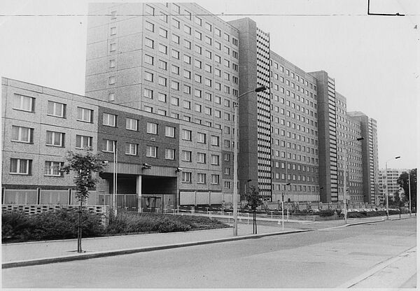 Schwarz-Weiß-Foto eines mehrgeschössigen Plattenbaus. Im Vordergrund ist eine nicht befahrene Straße.