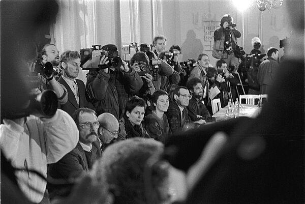 Mehrere Menschen sitzen nebeneinander an einem langen Tisch, vor, neben und hinter ihnen ist eine Vielzahl an Medienvertretern mit Kameras, Fotoapparaten etc. zu sehen.