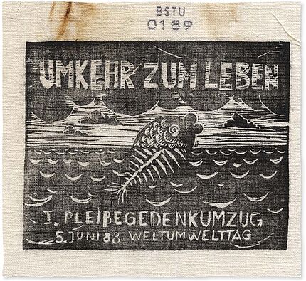Bedrucktes Stoffstück, auf dem ein Fisch abgebildet ist, der aus dem Wasser herausschaut. Der untere, sich im Wasser befindliche Teil des Fisches besthet nur aus Gräten. Darüber steht der Text "Umkehr zum Leben", darunter "1. Pleißegedenkumzug, 5. Juni '88, Weltumwelttag