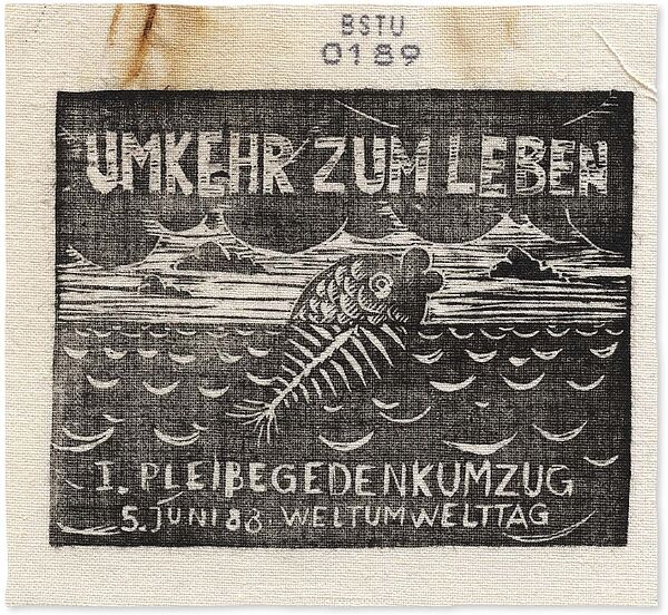 Schwarz bedrucktes Stoffstück, auf dem ein Fisch abgebildet ist, der aus dem Wasser herausschaut. Der untere, sich im Wasser befindliche Teil des Fisches besteht aus Gräten. Überschrift "Umkehr zum Leben"