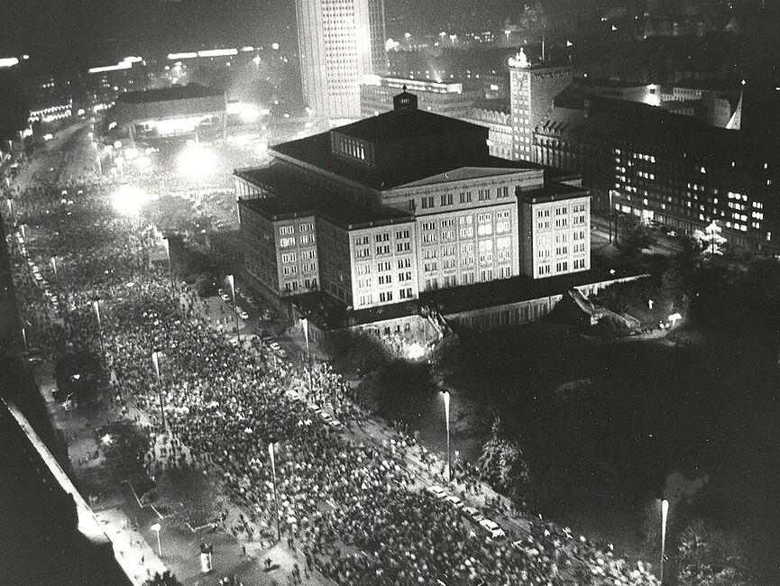 In der Bildmitte sieht man ein großes Gebäude, die Leipziger Oper. Auf der ringläufig davor verlaufenden großen Straße ist eine Menschenmenge versammelt.
