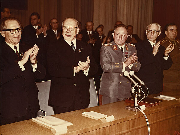 An einem Tisch stehen nebeinander aufgereits fünf freudig blickende Männer und klatschen in die Hände.