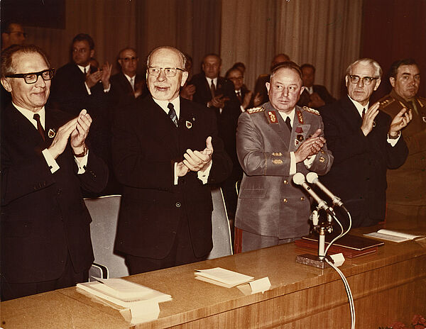 An einem Tisch stehen nebeinander aufgereits fünf freudig blickende Männer und klatschen in die Hände.