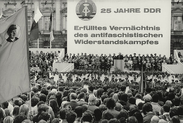 Blick auf das mit Blumen geschmückte Podium einer Kundgebung, auf dem zahlreiche Gäste sitzen. Ein Redner steht am Pult. Das Podium wird von einem Banner überragt. Es trägt neben dem Wappen der DDR den Text: "25 Jahre DDR. Erfülltes Vermächtnis des antifaschistischen Widerstandskampfes. Vor dem Podium sitzt eine Militärkapelle, davor drängen sich die Zuschauer. Im Vordergrund ist auf einer Fahne ein Portrait Ernst Thälmanns zu erkennen.