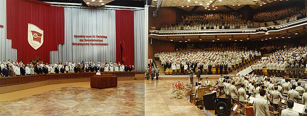 Aus zwei einzelnen Aufnahmen zusammengesetzes Foto im Querformat. Blick in den Veranstaltungssaal des Palasts der Republik. Links ein von zahlreichen Personen, zum Teil in weißen Gala-Uniformen der Stasi gekleidet, besetztes, sehr großes, geschmücktes Podium. Rechts gefüllte Zuschauerränge.