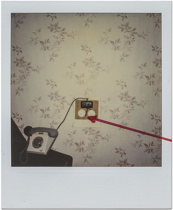 Manipulierte Telefonverteilerdose in einem Zimmer des Interhotels "Warnow" in Rostock, 22. Januar 1987; Quelle: BArch, MfS, BV Rostock, Abt. 26, Nr. 140, Bl. 2, Bild 2 Das Polaroidfoto zeigt eine tapezierte Wand, vor der ein Telefon steht. Neben dem Telefon ist eine elektronische Apparatur zu sehen. Diese wurde auf dem Polaroidfoto mit einem roten Pfeil markiert.