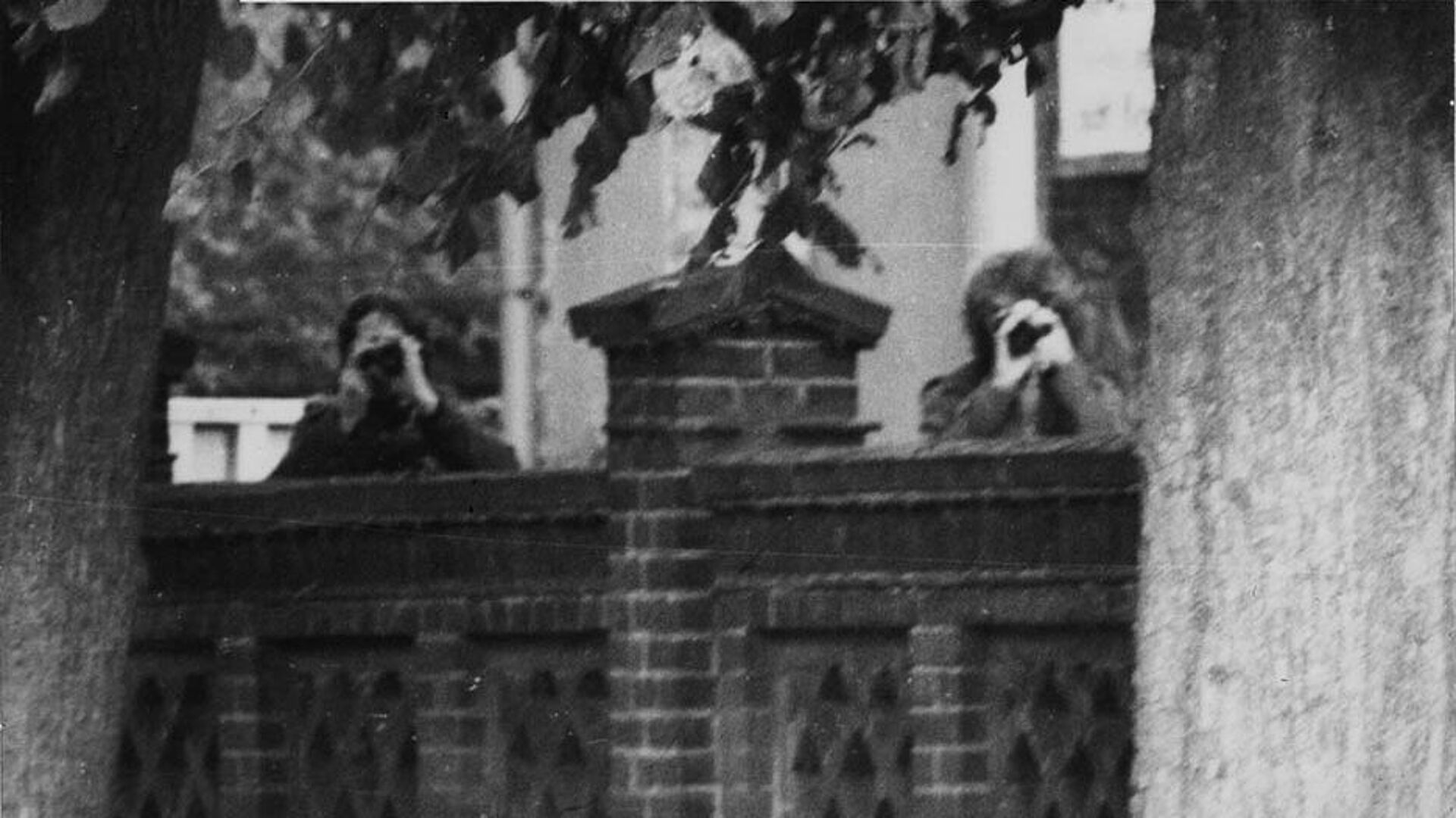 Schwarz-Weiß-Foto: zwei Männer schauen mit Ferngläsern über eine Mauer in Richtung des Fotografen.